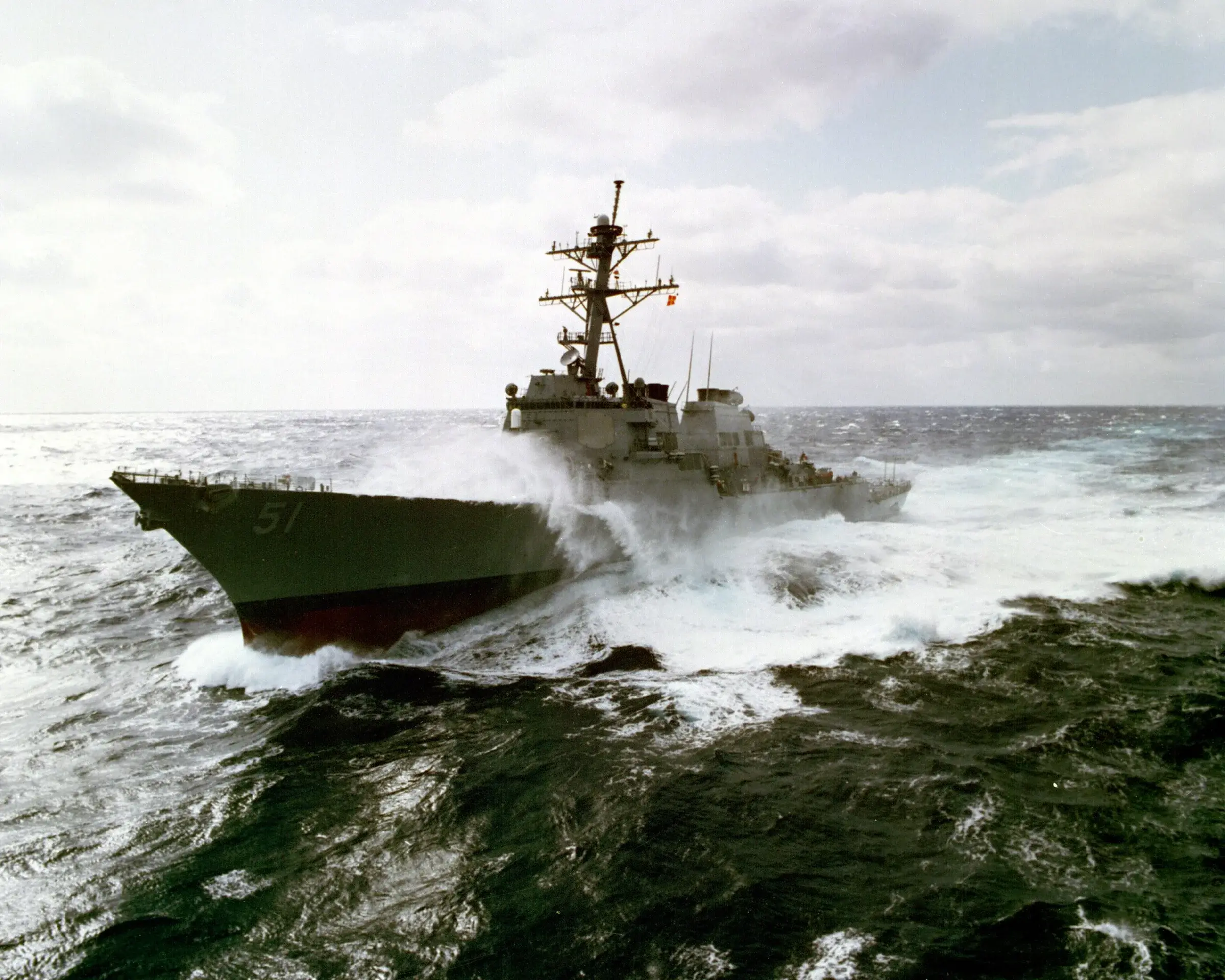 Le destroyer USS Arleigh Burke (DDG-51) navigue par mer formée, son mât chargé d'antennes et de radars visible au-dessus de la superstructure