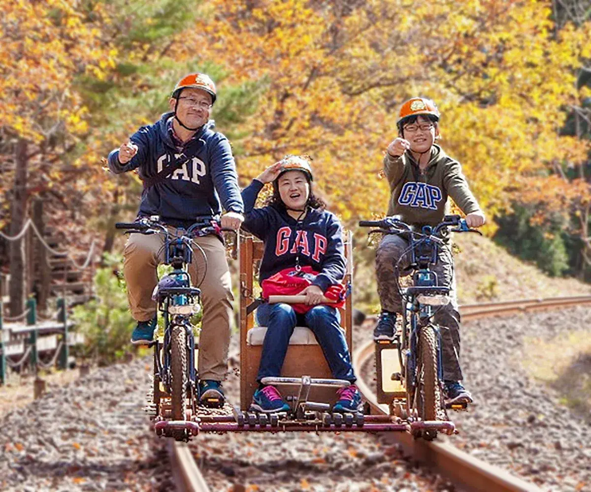 Famille sur un rail bike à banc central : deux pédaleurs et une passagère sur le siège bois, automne à Kamioka